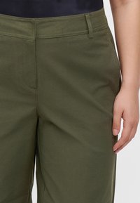 Personne portant un pantalon vert olive avec des passants de ceinture et une poche latérale visible, main reposant détendue à côté de la jambe.