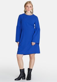 Blaues langärmliges Kleid mit Rundhalsausschnitt und ausgestellten Ärmeln, aus einem glatten Stoff gefertigt. Schwarze Absatzstiefel ergänzen das Outfit.