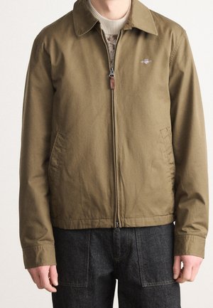 Homme portant une veste zippée vert olive sur un pull beige à motifs et un pantalon gris foncé, debout les mains relâchées le long du corps.