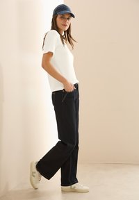 Bílý top s krátkým rukávem v kombinaci s tmavě modrými džíny. Model má na sobě námořnickou čepici a bílé tenisky se zelenými detaily, stojí na neutrálním pozadí.