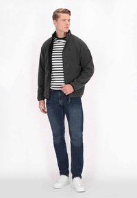 Giacca zip grigia scura, camicia a righe bianche e nere sotto, jeans blu e sneakers bianche. Texture lisce e design casual.