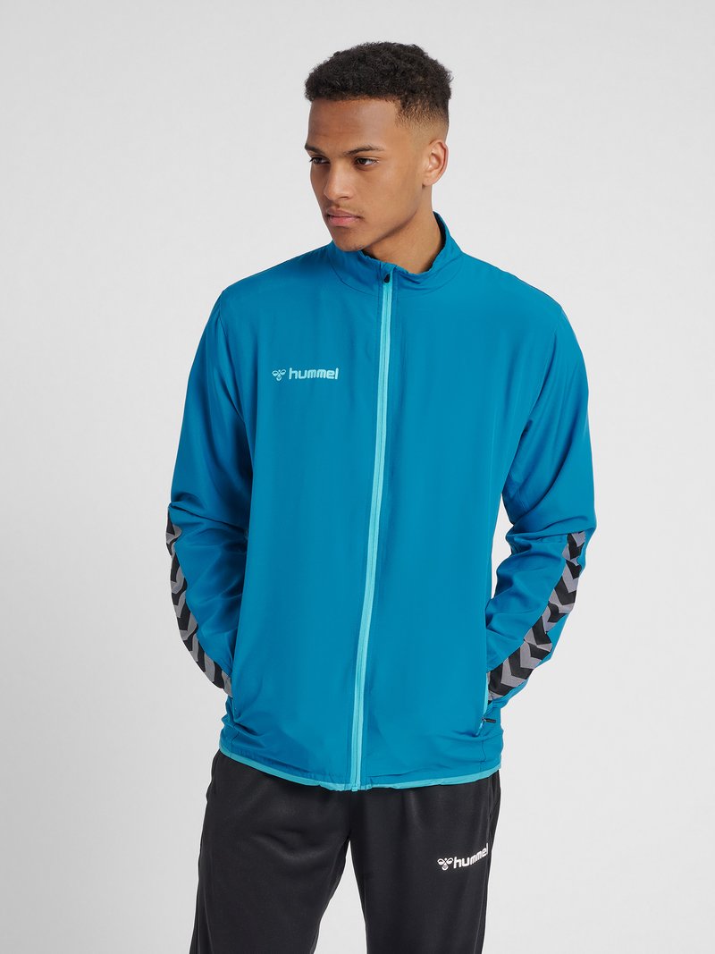 Turkooise lichtgewicht zip-up jack met een hoge kraag, voorzien van zwarte chevronaccenten op de mouwen en een logo op de borst.