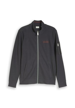 Chaqueta gris oscuro con cremallera completa y cuello alto, dos bolsillos frontales, bolsillo con cremallera en la manga y pequeño logo bordado en rojo en el pecho.