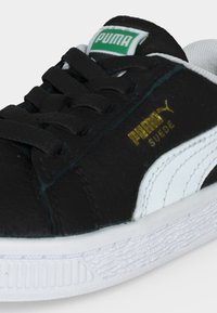 Baskets Puma Suede noires et blanches avec des lacets noirs, un logo vert sur la languette et un marquage doré Puma Suede sur le côté.