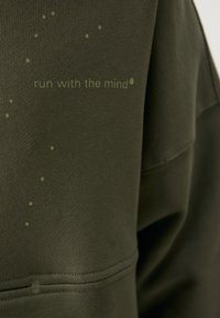 Olivengrøn sweatshirt med en struktureret overflade; har sætningen "løb med sindet" i lys grøn, udsmykket med små prikkede detaljer.