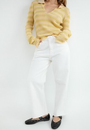Žena oblečená v žltom pruhovanom svetri, bielych nohaviciach s vysokým pásom a čiernych plochých topánkach stojaca na bielom pozadí.