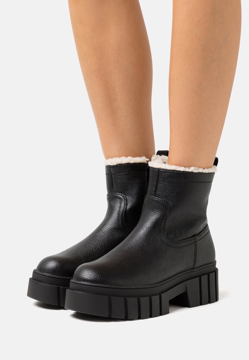 Bottines en cuir noir texturé avec semelles épaisses à rainures et doublure en polaire blanche, portées sur des jambes nues sur un fond clair uni.