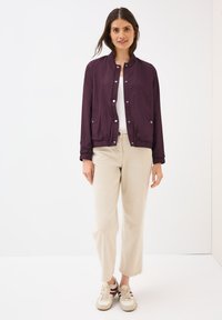 Een vrouw staat glimlachend met een paarse jas over een witte top, beige broek en sneakers. Ze poseert tegen een effen witte achtergrond.