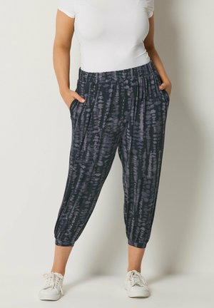 Kobieta ubrana w luźne, czarno-szare spodnie ¾ z wzorem tie-dye i kieszeniami, białą koszulkę oraz białe sneakersy na sznurowadła.