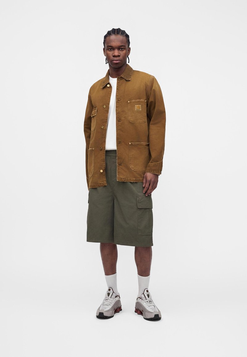 Uomo che indossa giacca marrone con bottoni, camicia bianca, pantaloncini cargo verde oliva, calzini bianchi e scarpe da ginnastica grigie, in piedi su sfondo bianco.