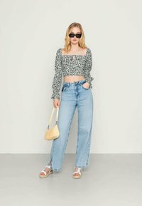 Jovem mulher vestindo um top cropped preto com flores, jeans de cintura alta azul clara com fendas frontais, sandálias brancas, óculos de sol pretos e segurando uma bolsa bege.