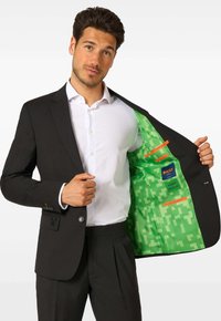 Čierny blazer s bielou košeľou pod ním, so žiarivou zelenou vzorovanou podšívkou a oranžovými akcentmi, zobrazený rozopnutý so štíhlym dizajnom.