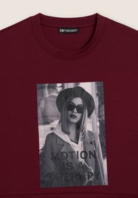 Donna con occhiali da sole e cappello in una foto in bianco e nero stampata su una maglietta color bordeaux con il testo "IL MOVIMENTO È UNA TERAPIA."