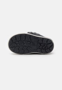 Suela de una zapatilla oscura, mostrando un patrón de tracción de goma con un borde claro. El fondo es completamente blanco, lo que resalta el calzado.