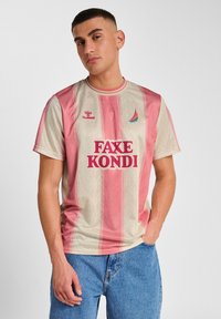 Krótki rękaw sportowej koszulki w jasnobeżowym kolorze z różowymi pionowymi paskami. Zawiera tekst "FAXE KOND" w grubym czerwonym kolorze oraz małe logo.