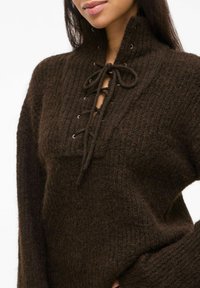Brauner, strukturierter Pullover mit hohem Kragen und Schnürdetails am Ausschnitt. Dickes Strickmaterial mit einem strukturierten Muster an den Ärmelbündchen.