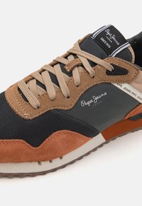 Zapatillas deportivas de malla negra con detalles en ante marrón. Cuentan con cordones beige y una suela de goma con una franja blanca en contraste.