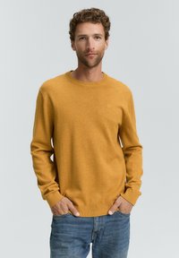 Suéter de punto color mostaza con mangas largas y cuello redondo. Textura suave, puños y dobladillo acanalados, con un pequeño logo en el pecho.