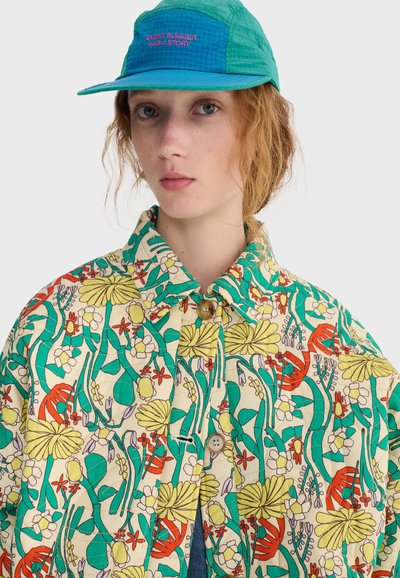 Jeune personne à la peau claire, cheveux roux bouclés, portant une casquette verte et bleue avec le texte "Chaque été a une histoire" et une veste colorée à motifs floraux.