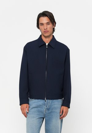 Chaqueta azul marino con cuello tipo solapa, cremallera delantera, dos bolsillos laterales y una textura de tela suave. Combinada con jeans de color azul claro.