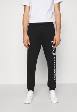Personne portant un pantalon de survêtement noir avec le logo blanc « EA7 Emporio Armani » sur la jambe droite, des baskets blanches et un t-shirt blanc uni à manches courtes.