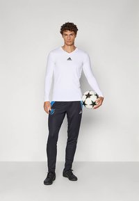 Férfi fehér Adidas hosszú ujjú pólóban és fekete Adidas melegítőnadrágban, fekete-fehér focilabdát tartva, egy sima világos háttér előtt áll.