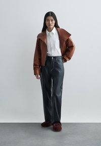 Giacca cropped in pelle marrone con ampio colletto, camicia bianca con bottoni, pantaloni a gamba larga neri e ciabatte pelose bordeaux. Sfondo semplice.