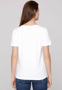 Camiseta blanca de mangas cortas hecha de una tela liviana y con textura irregular. Presenta un escote redondo y una pequeña impresión del logo cerca de la parte superior de la espalda.