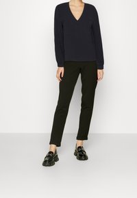 Długi rękaw, granatowa bluzka z dekoltem w serek w połączeniu z ciemnozielonymi spłaszczonymi spodniami i błyszczącymi czarnymi loafersami na platformie. Proste faktury, brak wyraźnych wzorów.