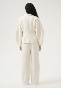 Kremno obarvana tailored jakna s nabranimi rokavi, v kombinaciji z širokimi hlačami. Teksturovana tkanina z gladko površino in strukturno silhueto.