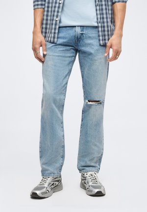 Homme portant un jean déchiré bleu clair, une chemise à carreaux, un t-shirt bleu clair et des baskets grises, debout sur un fond blanc.