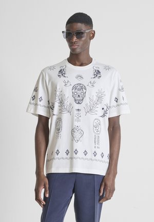 Uomo che indossa una maglietta bianca oversize con disegni in stile tatuaggio nero di teschi e fiori, abbinata a occhiali da sole scuri e pantaloni blu navy.