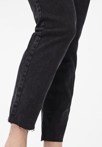 Parte inferior de las piernas con jeans de mezclilla negros con dobladillos deshilachados sobre un fondo blanco liso.