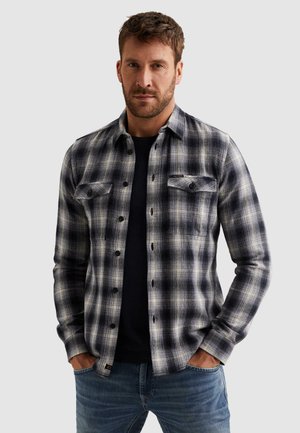 Mann mit kurzem braunem Haar und Bart, trägt ein schwarz-weiß kariertes Hemd über einem schwarzen Oberteil, steht mit den Händen in den Taschen.