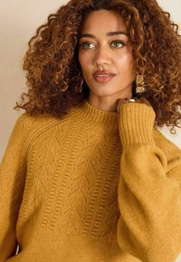 Mustár-sárga köt�ött pulóver, bordás nyakkal és cseh vonalakat ábrázoló textúrált mintával. A modell egy kézzel a nyaka mellett pózol.