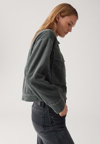 Gekort jasje van corduroy in donkergrijs met ribbelstructuur, voorzien van een knoopsluiting aan de voorkant en twee borstzakken. Gepaard met donkere jeans.