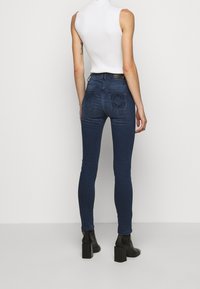 Mujer de pie, vistiendo jeans ajustados azul oscuro con bordado negro en los bolsillos traseros, un suéter sin mangas de cuello alto blanco y botas de tacón negras.