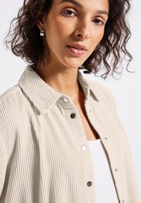 Vrouw met kort krullend haar, gekleed in een beige ribgebreide knoopjasje en zilveren creolen, kijkt naar de camera.