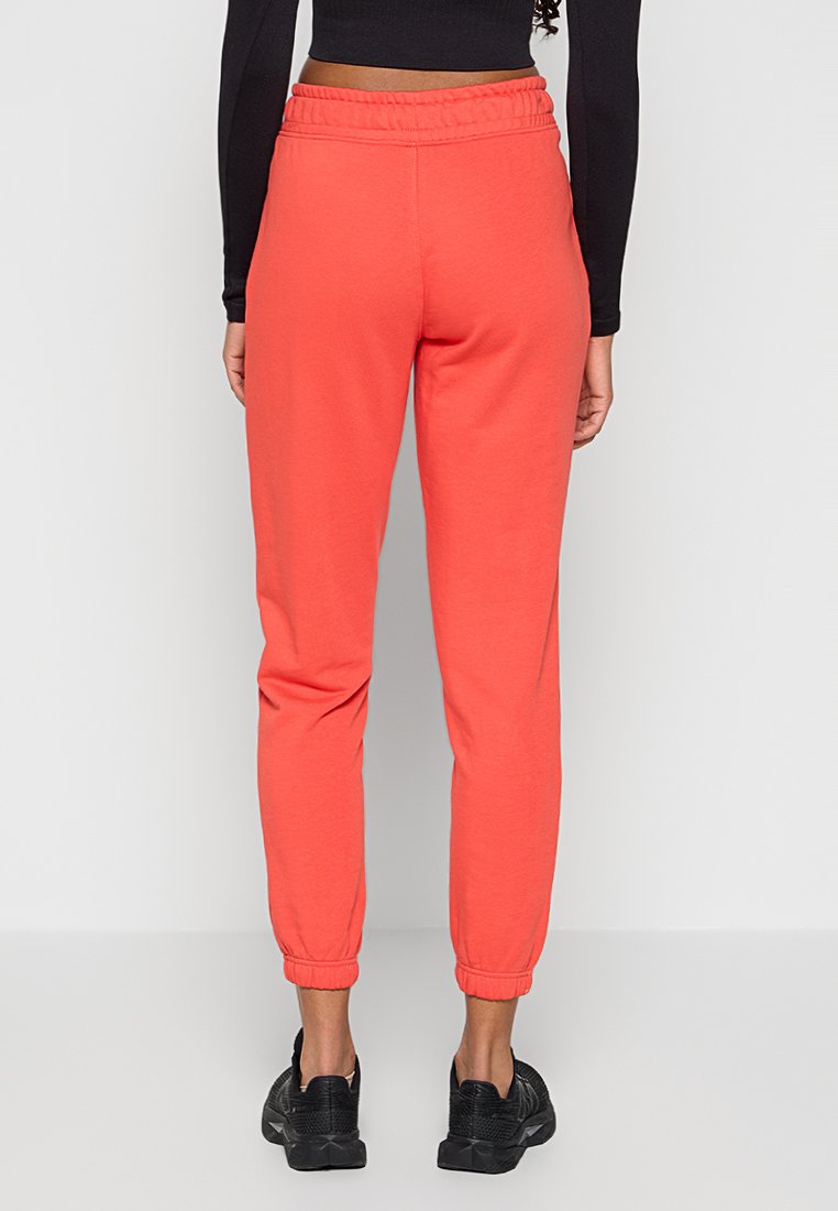 Frau in korallenfarbenen Jogginghosen mit elastischem Bund und Bündchen, einem schwarzen, langärmligen Cropped-Top und schwarzen Sportschuhen, die mit dem Rücken zur Kamera steht.