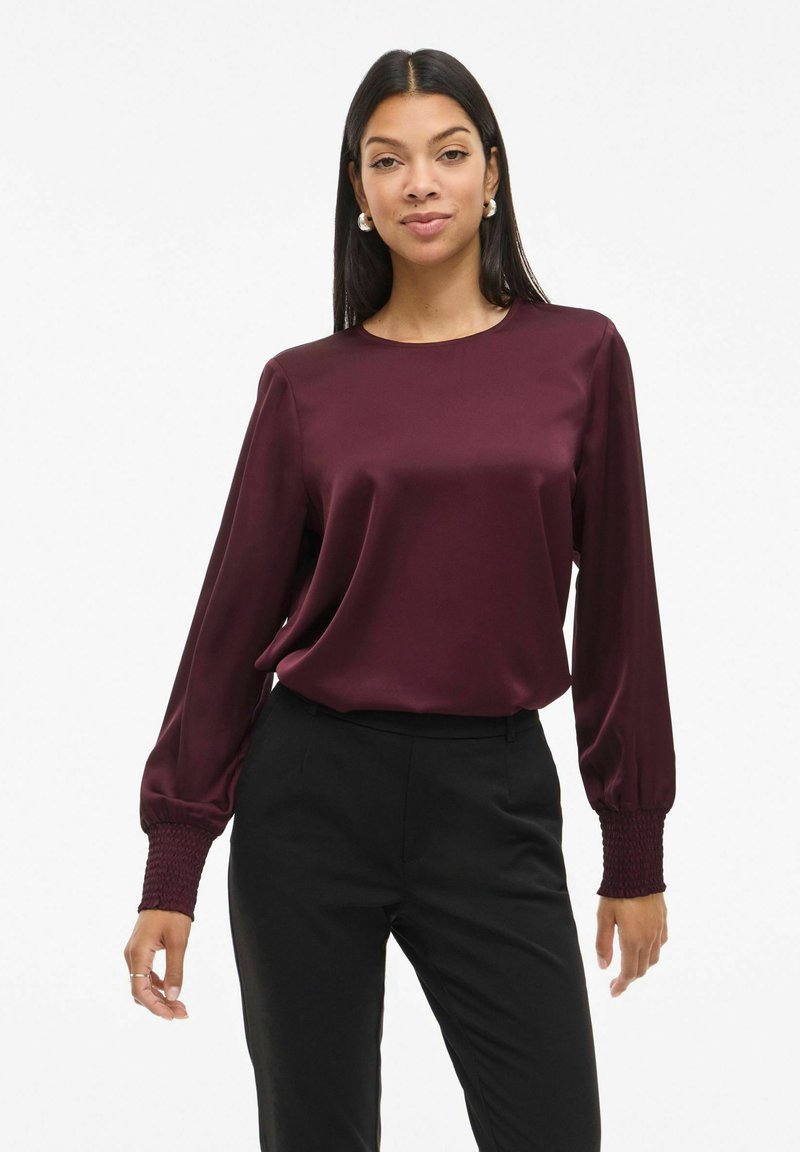 Femme portant un chemisier en satin bordeaux à manches longues avec poignets smockés et un pantalon noir, debout devant un fond blanc uni.