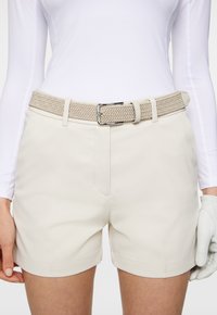 Beżowe szorty wykonane z gładkiego materiału, z bocznymi kieszeniami, złożonymi mankietami i plecionym beżowym paskiem z srebrną klamrą, zestawione z białą bluzką.