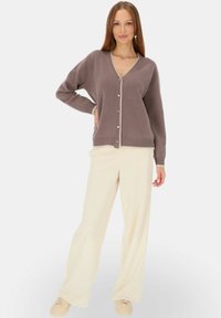 Brązowy kardigan z dekoltem w serek, zapinany na pięć guzików, z kontrastowymi wykończeniami w kolorze kremowym. Noszony na kremowych szerokich spodniach i jasnych butach.
