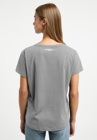 Een vrouw met lang bruin haar draagt een grijs T-shirt, waarbij het ontwerp aan de achterkant en de halslijn zichtbaar is, gecombineerd met lichtblauwe jeans tegen een effen achtergrond.