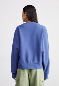 Modrý crewneck svetr s dlouhými rukávy a žebrovanými manžetami, nabízí volný střih a měkký bavlněný materiál. Mezi detaily patří prošívaný lem.