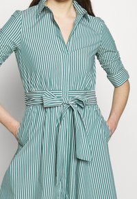 Abito camicia a righe turchesi e bianche con colletto, maniche a tre quarti, vita legata e tasche laterali. Realizzato in tessuto leggero.