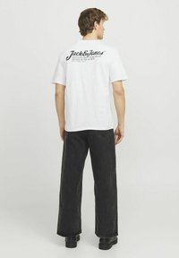 Camiseta blanca de algodón con texto impreso en negro en la parte posterior, combinada con pantalones anchos negros y zapatos negros, vista por detrás.