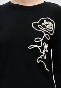 Pull noir en maille avec une corde blanche formant un motif floral abstrait sur le haut gauche de la poitrine.