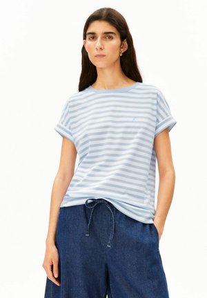 Jeune femme aux longs cheveux foncés portant une chemise rayée bleu clair et blanc et un pantalon bleu foncé à cordon sur fond blanc.