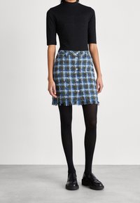 Silueta de falda a cuadros con flecos en el dobladillo, que presenta tonos azules y verdes, con botones en el frente, combinada con una parte de arriba ajustada negra y medias negras. Mocasines negros.