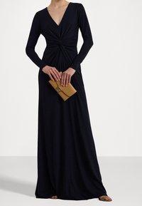 Lång svart klänning med djup V-ringning, långa ärmar och en tvistad detalj framtill. I kombination med en beige kuvertväska och enkla sandaler.